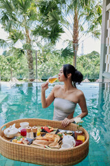 Happy woman in swimsuit having floating breakfast tray in luxury pool hotel. Young female enjoying sparklling wine in tropical resort. Relaxing, Exotic summer travel, holiday, vacation and weekend - 517535799