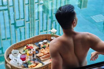 Happy man in swimsuit having floating breakfast tray in luxury pool hotel. Young male enjoying morning coffee in tropical resort. Relaxing, Exotic summer travel, holiday, vacation and weekend - obrazy, fototapety, plakaty