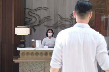 Hotel receptionist wearing medical mask as precaution against virus, welcoming and greeting a man who is walking to the desk to check in