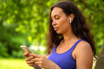 Beautiful girl stands in the park with wireless headphones and looks at the phone