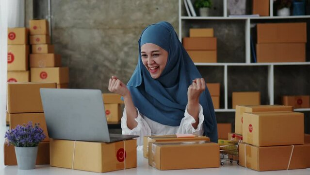 Female Muslim business monger  happy and cheerful for successful for receiving orders from customer via laptop with a lot of parcel boxes for shipping.
