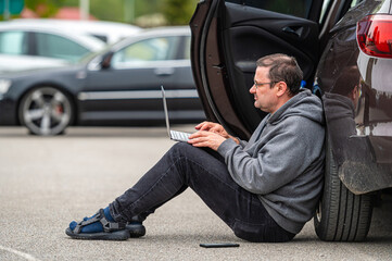 man sitting on asphalt by car and working on laptop, working remotely, traveling