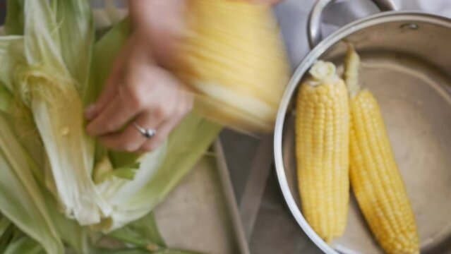 view from above, female hands with a ring on their finger and painted nails remove the peel from ripe corn cobs and put the peeled cobs in a pan for subsequent boiling in water.