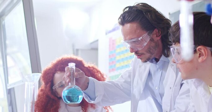 Close up of Caucasian young male teacher of chemitry subject working with pupils and showing chemical experiment with liquids in classroom. Children with man master in gogles learning at school.