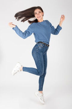 Beauty, fashion and lifestyle concept. Studio portrait of happy and beautiful woman with blue jeans, sweater and white sport shoes jumping in air studio portrait. White background with copy space