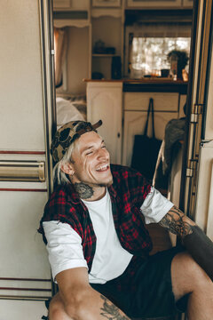 Hipster Looking Young Tattooed Dyed Blond Male In Red Plaid Sleeveless Flannel Dressed Upon White T-shirt, Wearing Camouflage Cap, Earrings And Black Shorts Chilling Near Opened Door Of His Camper