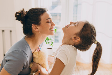 Adorable smiling female in gray clothes with hair bun reaching her face to kiss cheerful daughter,...