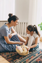 Pretty babysitter in gray casual dress telling fairy tale to little curious girl in denim skirt and white blouse sitting on vintage carpet with plush light brown bear. Human relationships