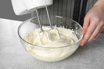 Woman whipping white cream with mixer at light grey table, closeup