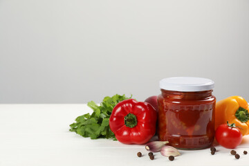 Glass jar of delicious canned lecho and fresh ingredients on white wooden table. Space for text