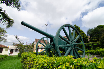 canhão antigo na cor verde em praça com céu azul com nuvens e casa colonial
