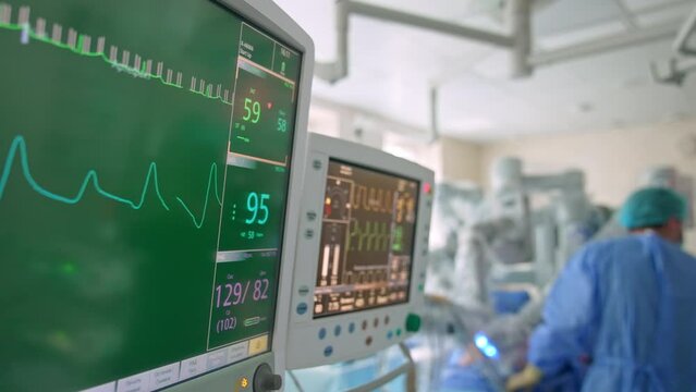 Two screens showing various life parameters of a patient at operation. Doctors standing near Da Vinci robot at backdrop in blur.