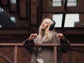 Woman wearing headphones. Young and stylish woman with long blonde hair wearing beige coat listening music with smartphone and wireless  headphones. Music, social media, lifestyle concept.