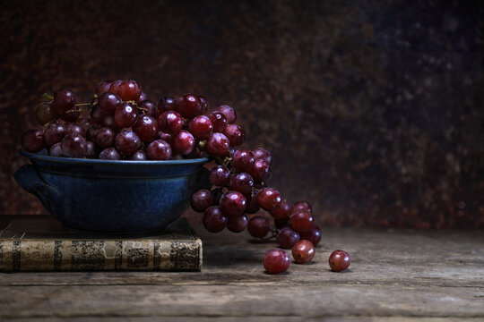 Fresh red grape in a blue bowl with a vintage bood on wooden board table and background - dark and moody