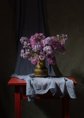 A large bouquet of lilacs in a green earthen jug. Green vase on the table. Dark background. Blue curtain. The scent of spring. Peace of mind. Comfort at home. Joyful mood. Cheerful still life.