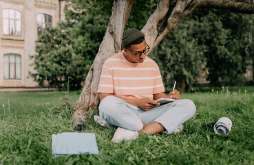 Happy african american guy writing something in notebook, sitting in park, studying outdoors and preparing for lecture