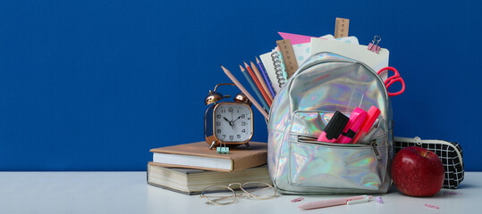School backpack with stationery, clock and apple on table against blue background