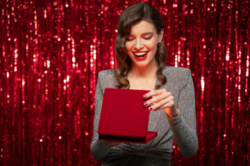 Happy surprised woman in party dress opening present box on red glittering tinsel background