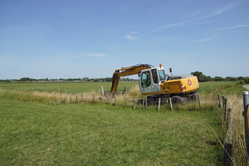 Dutch yellow excavator in a Dutch meadow landscape with ditch and trees in the distance. Summer, July, Netherlands                                                            