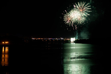 Huge colorful fireworks in the distance, on the shore of a city lake or river, with green, pink and...