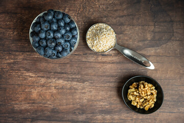 Glass goblet of fresh, nutritious, tasty blueberries, small bowl of walnuts, and a measuring cup with raw steel-cut oats on a rustic wood background

