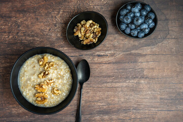 Healthy breakfast of cooked oatmeal in a black bowl, with fresh walnuts and blueberries, on a rustic wood background
