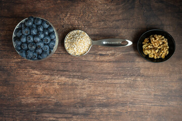 Glass goblet of fresh, nutritious, tasty blueberries, small bowl of walnuts, and a measuring cup with raw steel-cut oats on a rustic wood background
