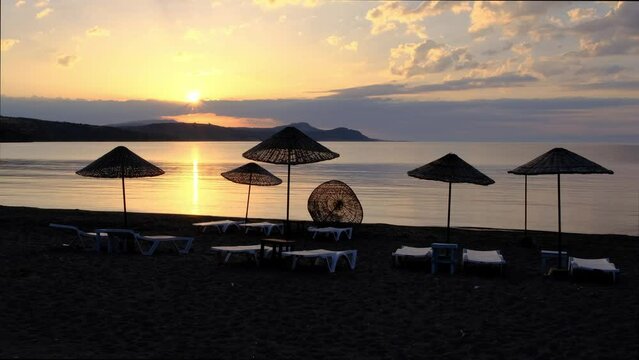 Sunrise over Aegean Sea, beach parasols and lounge chairs in the front. Calm flat water and dramatic sky.