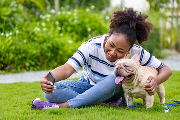 African American woman is taking selfie with her french bulldog puppy while lying down in the grass lawn after having morning exercise in the park.