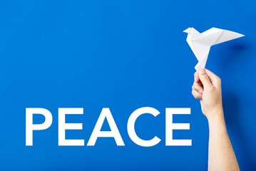 Word Peace in white letters on a blue background next to a hand holding a white paper dove, symbol...