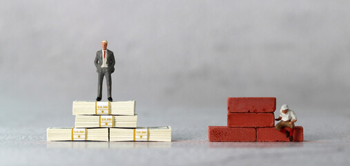 A miniature man standing on a $100 bill and a miniature man sitting on a red brick. The concept of...
