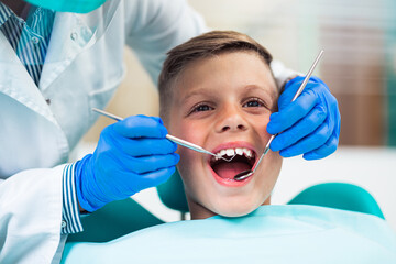 Cute little boy sitting on dental chair and having dental treatment. Dentist is wearing protective face mask due to Coronavirus pandemic.