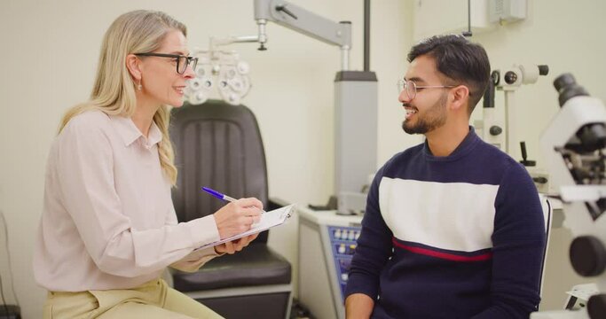 Mature optometrist talking, writing on clipboard in patient consult in an eye clinic. Ophthalmologist and professional optician explaining glaucoma or disorders to young man and listing prescription