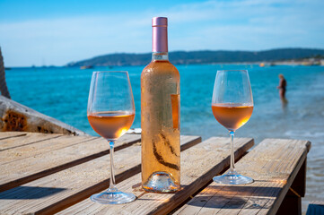 Glasses and bottle of cold rose wine from Provence served outdoor on wooden yacht pier with view on blue water and white sandy beach Plage de Pampelonne near Saint-Tropez, France