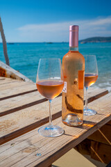 Glasses and bottle of cold rose wine from Provence served outdoor on wooden yacht pier with view on blue water and white sandy beach Plage de Pampelonne near Saint-Tropez, France