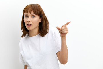 a sweet, emotional, playful woman in a white T-shirt stands on a white background and points with her index finger towards an empty space