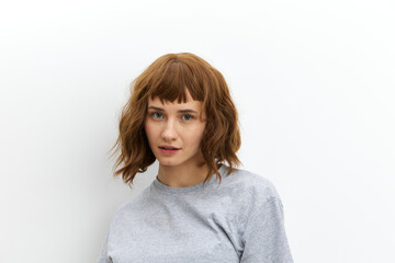 Obraz premium a sweet, inspired, beautiful red-haired, curly-haired woman stands on a white background in a gray cotton T-shirt and looks at the camera with a gentle gaze
