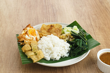 Nasi Pecel, a traditional Javanese rice with vegetable salad and peanut sauce, served with side dishes such as egg, martabak, and peyek crackers.
