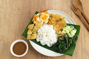 Nasi Pecel, a traditional Javanese rice with vegetable salad and peanut sauce, served with side dishes such as egg, martabak, and peyek crackers.
