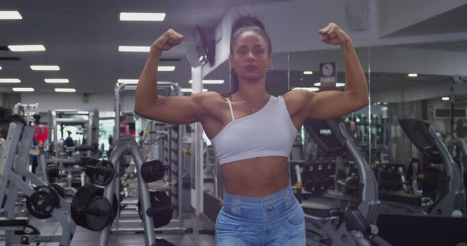 A strong fit muscle biulder flexing her muscles in a gym with eqiupment in the background