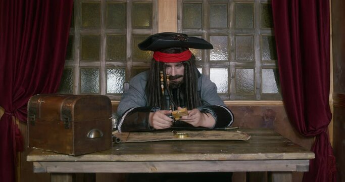 Pirate in hat and wig with dreadlocks sits down at table with treasure map and looks around with bored look. He takes small music box and winds it up to listen to uncomplicated melody