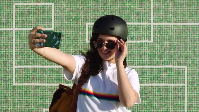 A cool female skater taking a selfie using her phone in a skate park on a windy summer afternoon. A trendy and stylish woman student snaps a picture of herself to share on social media