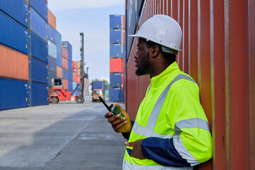 African American male worker in safety uniform and hardhat use walkie-talkie, work at logistics terminal with many stacks of containers, loading control shipping goods, cargo transportation industry.