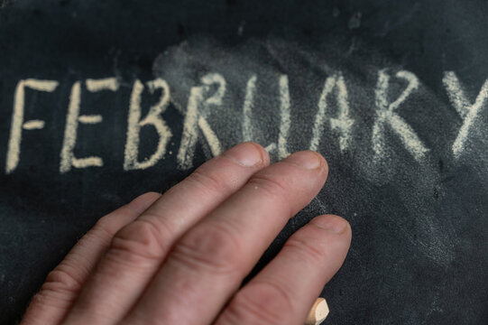 The semi-erased word FEBRUARY on the black chalkboard. An adult man's left hand removes the handwritten word with his fingers. Fuzzy letters on black surface. End of calendar month concept - Powered by Adobe
