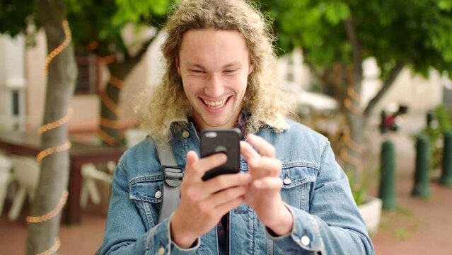 A stylish male student texting on his phone smiling and laughing at social media news. A young man browsing the internet and seeing clickbait using his cellphone outdoors in a park while traveling