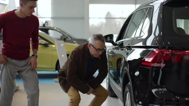 Father, son are examining new car and sitting on floor in dealership office spbd. 4k Close view of two caucasian men study transport and discuss details, make decision and sit in sales room. 2 family