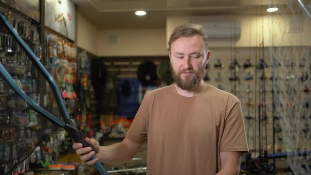 young bearded caucasian male fisherman in a fish tackle shop chooses a landing net for fishing, compares two options and buys the best one.