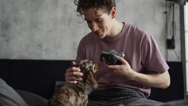 Young man is taking photo and touching dog while sitting on bed at home spbd. 4k Close view of handsome european guy holds camera and takes pictures of purebred pet, touches gently and looks with