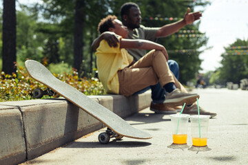 Horizontal image of skateboard and cold drinks standing on the ground with dad and son resting together in background