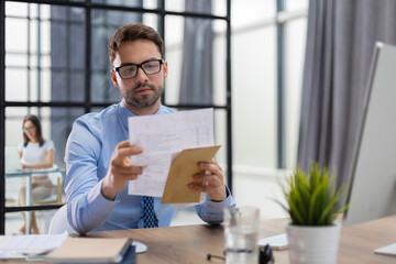 Handsome entrepreneur reading a letter from envelope in a desktop at office with collegues on the background.